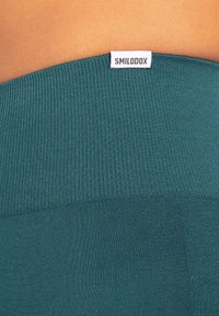 Ceinture côtelée teal des leggings avec une étiquette blanche "SMILODOX". La texture semble lisse et extensible, idéale pour les vêtements de sport.