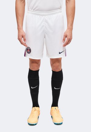 Muž oblečený v bielych futbalových šortkách Paris Saint-Germain, čiernych Nike podkolienkach a žltých kopačkách stojaci na jednoduchom pozadí.