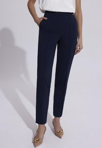Granatowe, dopasowane spodnie o wąskim kroju, wyposażone w boczne kieszenie i gładką teksturę, zestawione z beżowymi, szpiczastymi butami z złotymi akcentami.