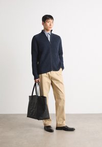 Suéter de cremallera acanalado en azul marino, pantalones beige, zapatos de cuero negros y un bolso tote negro con el logotipo "Hugo" en relieve.