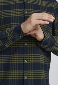 Marineblå og olivenruter button-up skjorte med en avslappet passform. Har grå knapper og justerbare mansjetter. Stoffet ser strukturert og pustende ut.