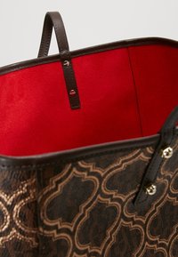 Brązowa torba na ramię z wzorem, z fakturowaną powierzchnią zewnętrzną, czerwoną podszewką wewnętrzną, skórzanymi uchwytami oraz złotymi elementami ozdobnymi.
