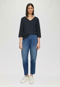 Maglione blu navy a V con maniche a tre quarti, abbinato a jeans in denim blu a vita alta e sneaker bianche con suola spessa. Sfondo neutro.