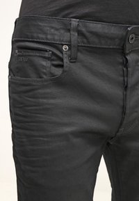 Jeans de mezclilla negra de corte ajustado, con bolsillo delantero, detalles en el bolsillo trasero, herrajes metálicos y textura sutil.