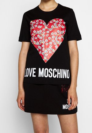 Vrouw draagt een zwart T-shirt en rok; het T-shirt heeft een groot rood hart met witte bloemen en de tekst ‘LOVE MOSCHINO’ in wit onder het hart.