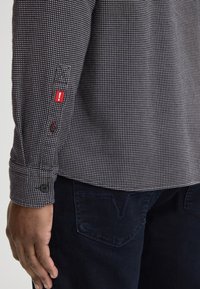 Donkerblauwe shirt met een klein wit geruit patroon. Heeft zwarte knopen en een rode tag op de mouw. Draagt met donkergekleurde broek.