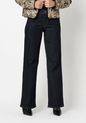 Woman wearing dark blue wide-leg jeans, paisley patterned jacket, and black pointed-toe boots, standing against white background.