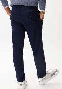 Granatowe spodnie o luźnym kroju, wykonane z gładkiej tkaniny. Posiadają tylne kieszenie zapinane na guziki i są noszone z białymi i szarymi sneakersami.