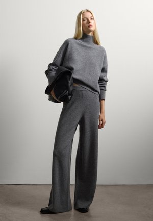 Pullover grigio in misto lana con collo alto e bordo a costine, abbinato a pantaloni grigi a gamba larga. Il modello tiene in mano una borsa nera con texture.