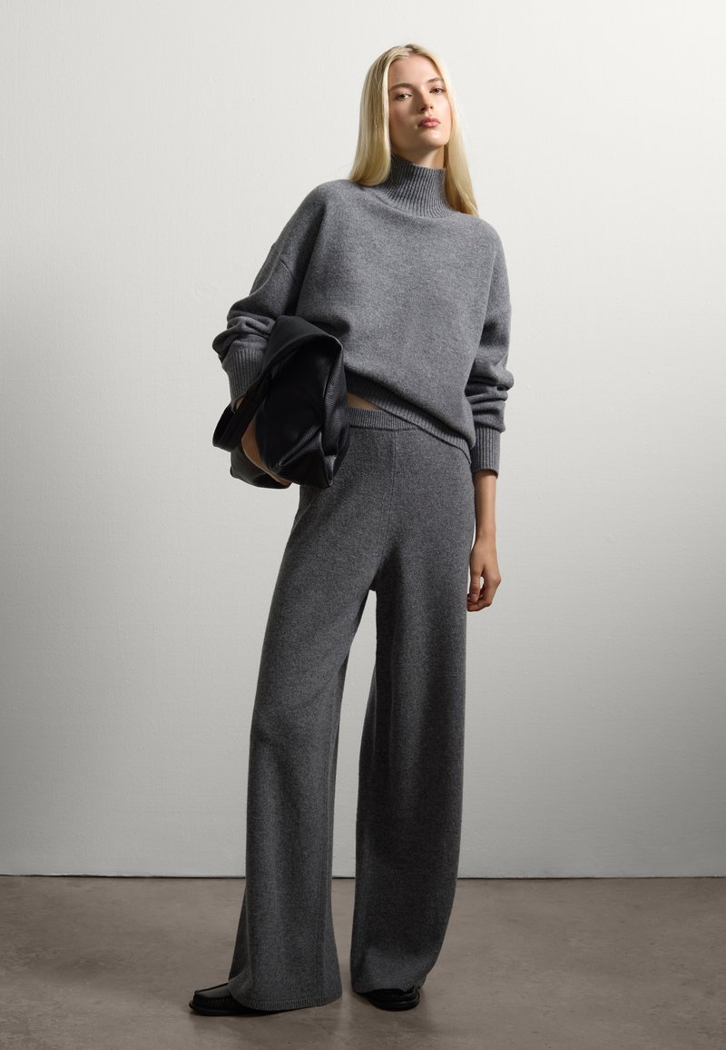 Pullover grigio in misto lana con collo alto e bordo a costine, abbinato a pantaloni grigi a gamba larga. Il modello tiene in mano una borsa nera con texture.