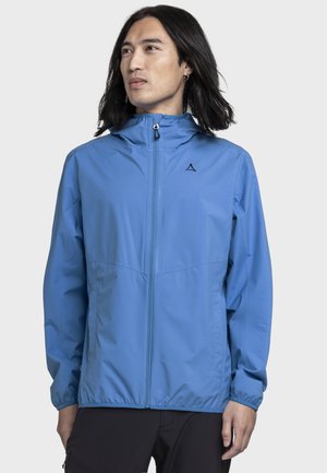 Person trägt eine langärmelige hellblaue Kapuzenjacke mit Reißverschluss, steht vor einem einfachen hellen Hintergrund und schaut zur Seite.