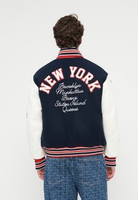 Chaqueta varsity en azul marino y blanco con "NEW YORK" en letras rojas, con nombres de ciudades bordados abajo. Cuello y puños de canalé, tela texturizada.