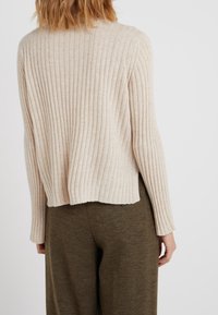 Jersey beige con fondo acanalado y mangas largas, que presenta un escote redondo y una parte trasera ligeramente recortada. Combinado con pantalones verde oliva.