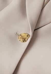 Tissu de blazer beige avec un seul bouton doré brillant de forme irrégulière fixé près du revers.