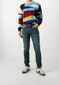 Färgglad stickad tröja med abstrakta mönster, bärs med blå jeans och flerfärgade sneakers med grå och neonkonturer.