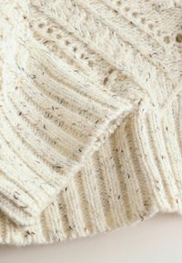 Jasna kremowa sweter w robótki na drutach ma prążkowaną strukturę z drobnymi plamkami ciemniejszych kolorów, prezentując warstwowy design i miękką, grubą włóczkę.