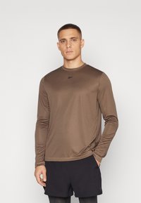 Brun langermet treningstrøye laget av glatt stoff, med rund hals og en liten logo på brystet. Kombinert med svarte shorts.