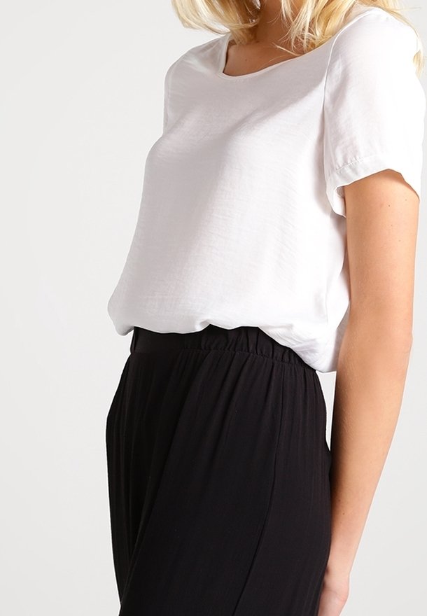 Femme portant un chemisier blanc uni à manches courtes, rentré dans un pantalon noir taille haute avec une ceinture élastique.