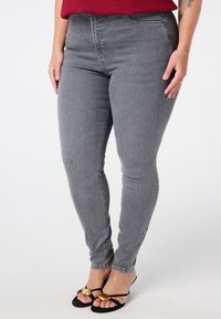 Jeans skinny gris en denim, avec une taille haute, des poches avant et une texture lisse ; associés à des sandales noires avec des accents dorés.