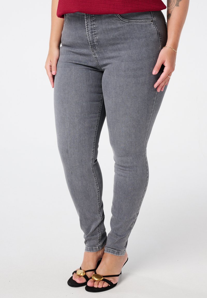 Jeans skinny gris en denim, avec une taille haute, des poches avant et une texture lisse ; associés à des sandales noires avec des accents dorés.