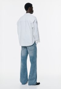 Cămașă albă cu mâneci lungi și blugi evazați de culoare albastră deschisă, având o textură moale a țesăturii și un croi clasic, cu buzunare vizibile pe spate.