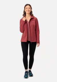 Giacca softshell rossa con colletto alto, indossata sopra una maglietta leggera, abbinata a leggings neri e scarpe da ginnastica blu, con logo riflettente.