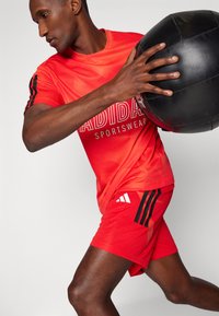 Rdeč športni komplet z mrežno teksturo; vključuje majico s kratkimi rokavi in kratke hlače, ki imata črne Adidas stripe in logotip.