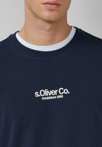 Marineblaues Sweatshirt mit Rundhalsausschnitt und geripptem Kragen, ausgestattet mit einem weißen Logo-Textdruck, der "s.Oliver Co. Gegründet 1969." liest.
