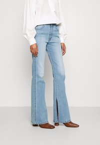 Frau in hellblauen Schlagjeans mit vorderen Schlitzen, kombiniert mit braunen spitzen Stiefeln und einer weißen Bluse mit langen Ärmeln.