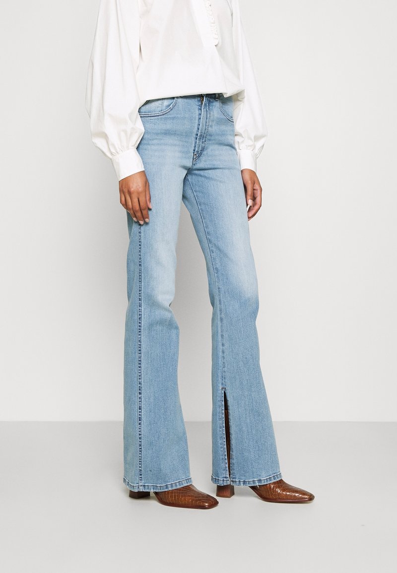 Frau in hellblauen Schlagjeans mit vorderen Schlitzen, kombiniert mit braunen spitzen Stiefeln und einer weißen Bluse mit langen Ärmeln.