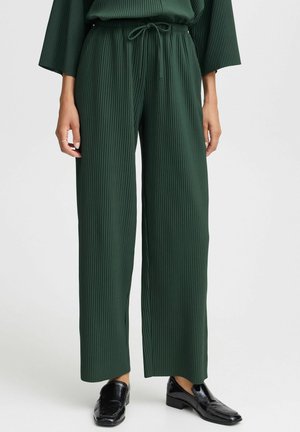 Pantaloni a gamba larga a coste verdi con cintura elasticizzata e cordino, che mostrano un tessuto testurizzato e una silhouette fluida. Abbinati a scarpe nere.