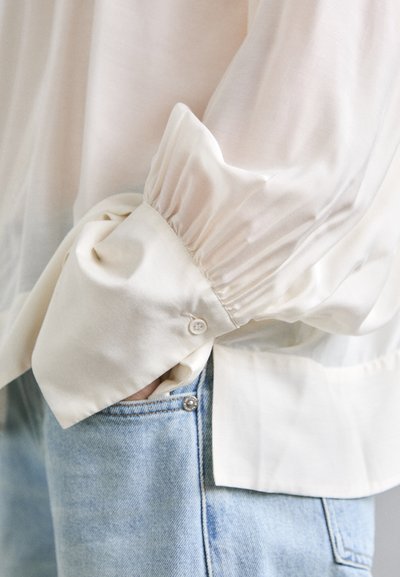 Blusa color crema chiara con maniche a sbuffo, dettagli con polsini arricciati con bottone, indossata sopra jeans in denim blu chiaro con vestibilità classica.