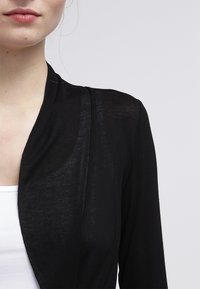 Černý lehký kardigan s přiléhavým střihem a otevřeným předním dílem. Vyrobený z průhledné tkaniny, s jednoduchou, bezšvovou strukturou a podrobným prošíváním.