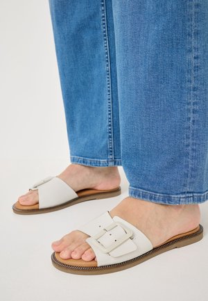 Personne portant un jean bleu et des sandales blanches à enfiler avec une grande boucle, debout sur une surface claire.