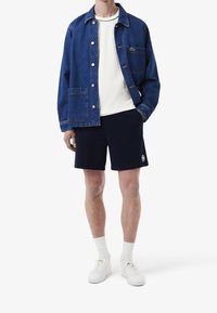 Giacca di jeans blu scuro con una tasca sul petto, abbinata a una maglietta bianca a girocollo, pantaloni corti blu navy e sneakers bianche, completate con calze bianche.