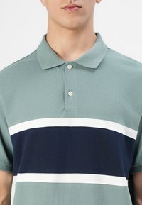 Moški nosi polo majico s kratkimi rokavi v zeleni barvi, ki ima dve beli vodoravni črti in široko mornarsko modro črto čez prsi.
