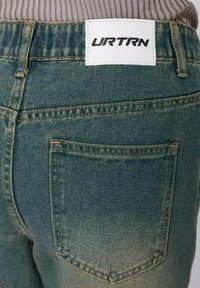 Μπλε τζιν παντελόνι με μεσαία μέση, με λευκή ετικέτα που γράφει "URTRN", και δύο πίσω τσέπες με κανονικό ράψιμο.