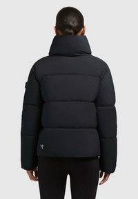 Persona con capelli scuri legati indietro, che indossa un giubbotto imbottito nero con colletto alto, vista di spalle su uno sfondo semplice.