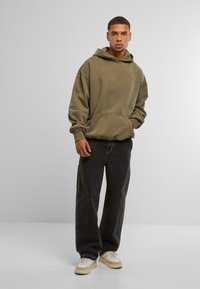 Olīvu zaļš džemperis ar priekšējo kabatu, brīvs piegriezums un ribota maliņa, kombinēts ar tumšām džinsiem un gaišiem sporta apaviem.