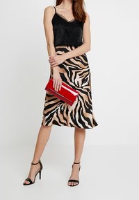 Camisola nera con rifiniture in pizzo, gonna a stampa animale nei toni beige e nero, pochette rossa, scarpe con tacco nere con cinturini, in piedi su uno sfondo neutro.