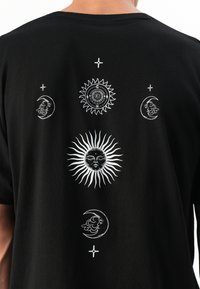 Schwarzes T-Shirt mit weißen himmlischen Designs, darunter eine große Sonne mit Gesicht, drei Halbmonde mit Gesichtern und kleinen Sternen.
