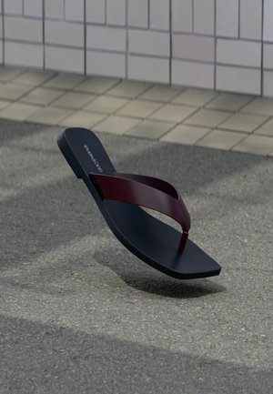 Ein einzelner schwarzer Flip-Flop mit einem dunkelroten Riemen, der über einer grauen Asphaltfläche schwebt, mit beigem gepflasterten Bürgersteig im Hintergrund.
