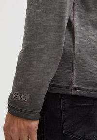 Langärmliges graues Shirt aus strukturiertem Stoff, mit roten Nähten und einem Camp David-Label auf dem linken Ärmel. Kombiniert mit dunklen Jeans.