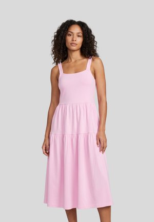 Une femme aux cheveux foncés et bouclés porte une robe midi rose sans manches à volants, sur un fond gris uni.