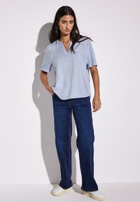 Blusa azzurra chiara a maniche corte con scollo a V e risvolti, abbinata a jeans a zampa d'elefante blu scuro e sneakers bianche. Tessuto liscio in tutto il capo.