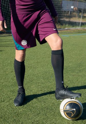 Zwarte voetbalschoenen op een voet die rust op een zwart-wit voetbal; de atleet draagt bordeauxrode shorts met een teal accent en zwarte kniehoge sokken.