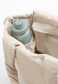 Beige quiltet taske med en lysblå vandflaske indeni. Tasken har en snorelukning i toppen og en lynlåslomme på siden.