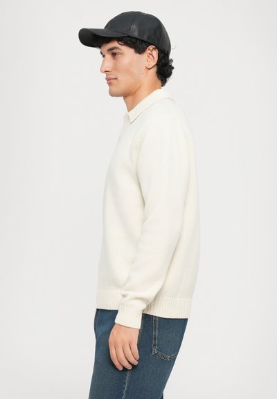 Suéter color crema con puños y dobladillo acanalados, llevado sobre unos jeans de mezclilla. Se lleva una gorra de cuero negra, añadiendo un elemento de contraste.