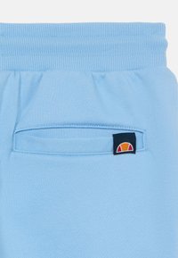 Lyseblå joggebukser har en ribbekant i livet, en liten baklomme og et merke med logo i røde, oransje og gule detaljer.