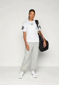 T-shirt branca da Adidas com detalhes em preto nas três riscas, combinada com calças de moletom cinzentas e sapatilhas brancas. Mochila preta pendurada no ombro.